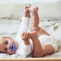 Niemowlę w białym body leży na plecach na łóżku, trzymając się za stopy.