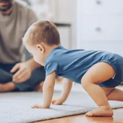 Dziecko w niebieskim body raczkuje na dywanie, a w tle siedzi ojciec.