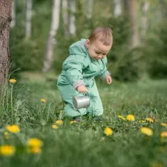 Dziecko w zielonym kombinezonie z konewką w trawie z żółtymi kwiatami.