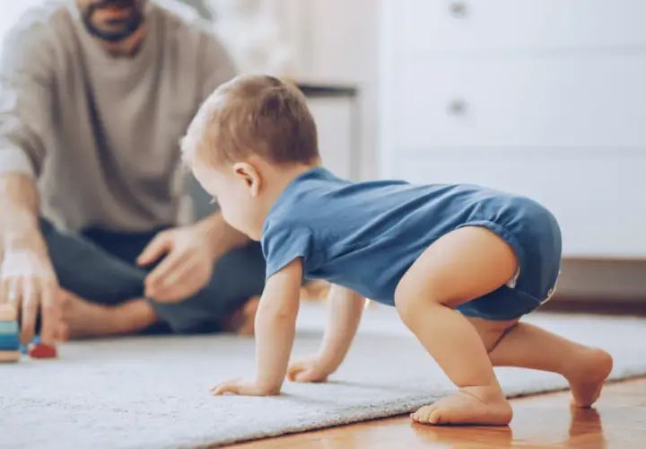 Dziecko w niebieskim body raczkuje na dywanie, a w tle siedzi ojciec.