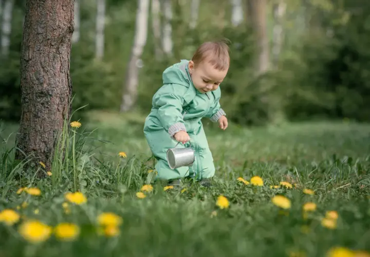 Dziecko w zielonym kombinezonie z konewką w trawie z żółtymi kwiatami.